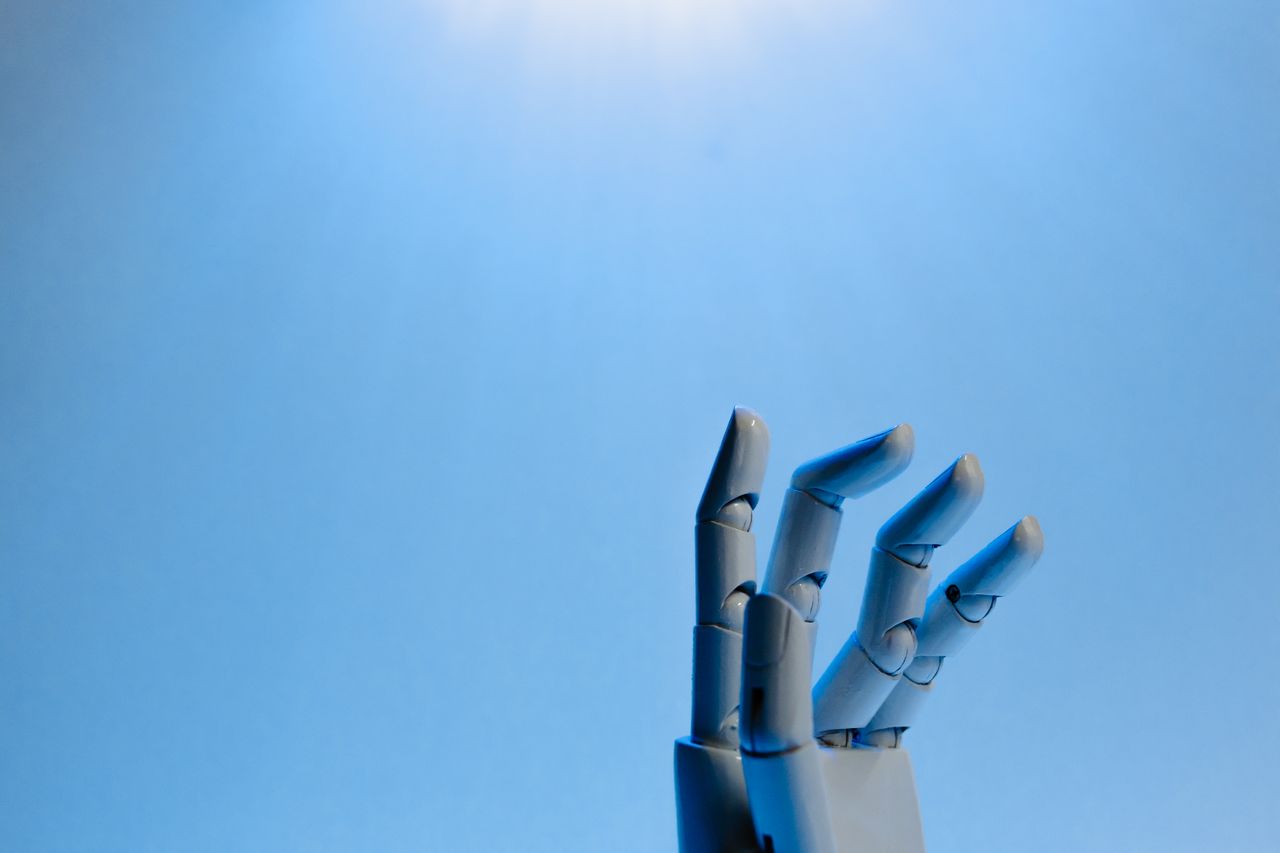 hero-img-01 Robotic hand with articulated fingers reaching towards the sky on a blue background.